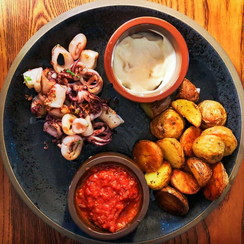 A black plate with seared calamari rings, roasted small potatoes, and two dipping sauces