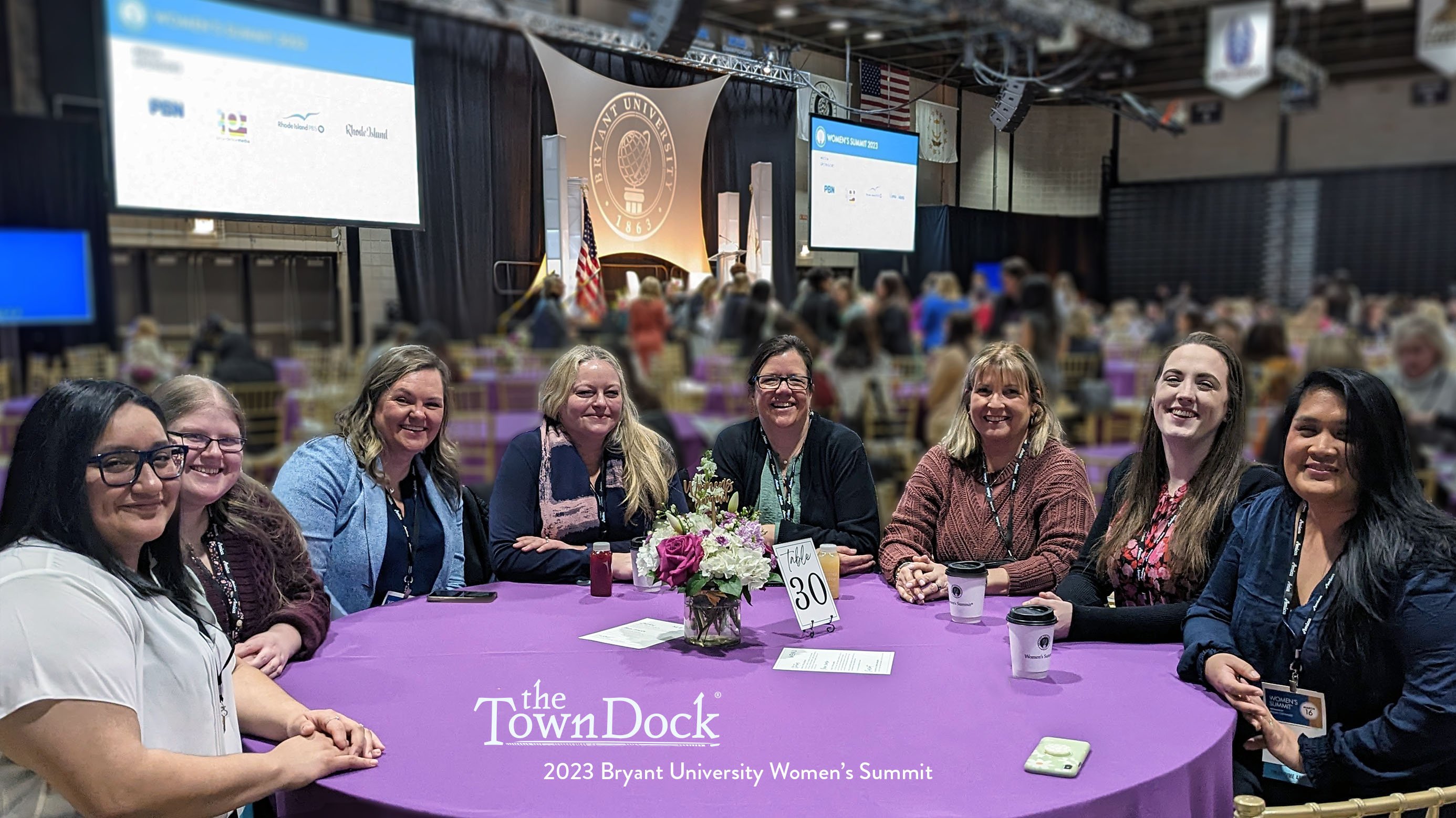 A group of women team members from The Town Dock sit around a purple table. The Bryant University mainstage is in the background.