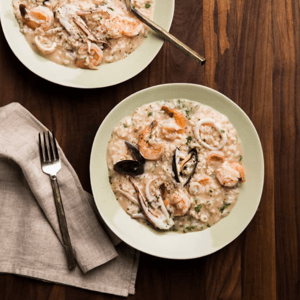 A light green bowl of risotto with calamari rings and other seafood on a dark wood table