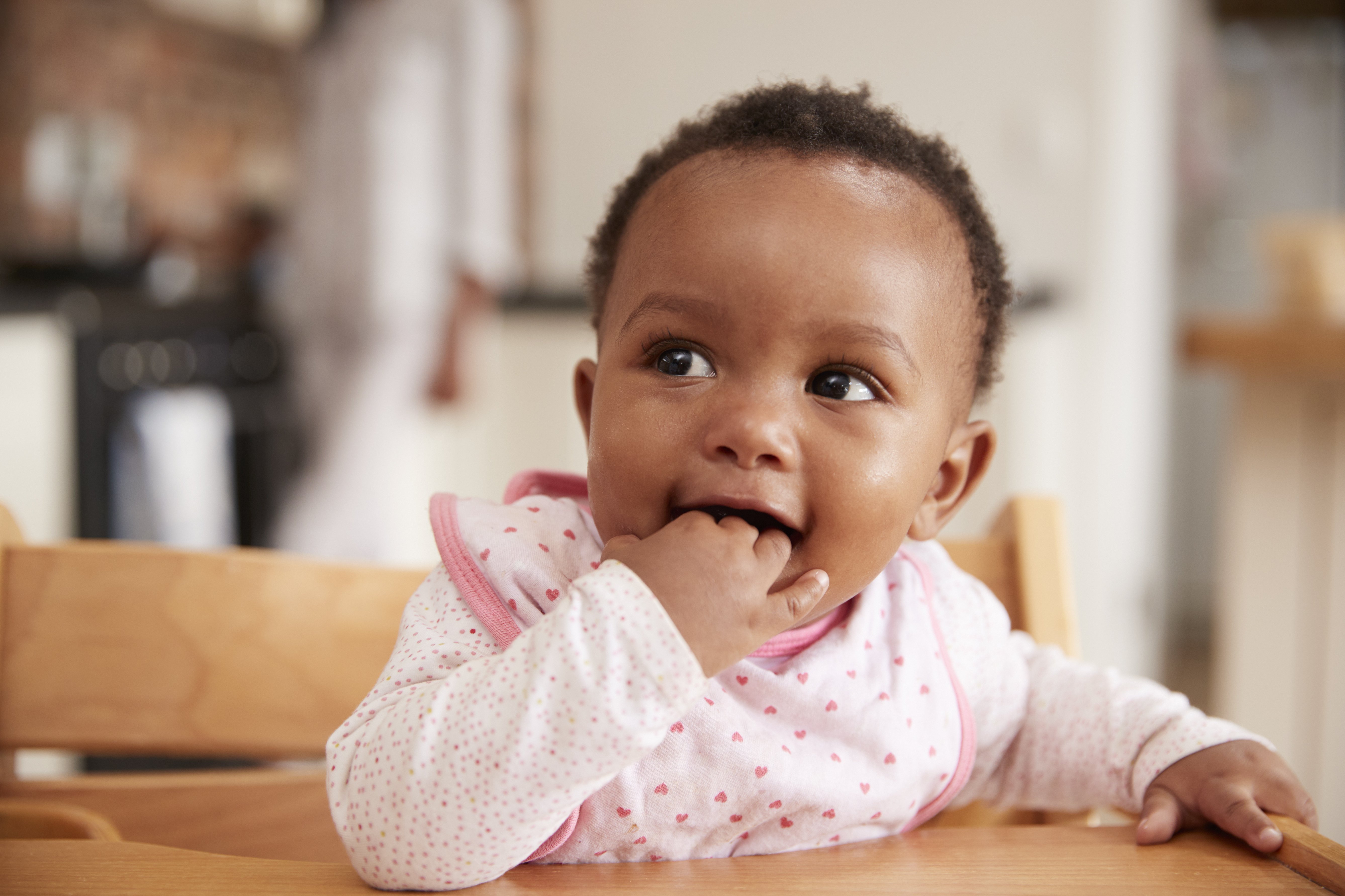 A Black baby dressed in pink heart bib sits at their highchair with fingers in mouth