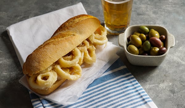 A baguette with fried calamari rings, a beer, and small dish of olies