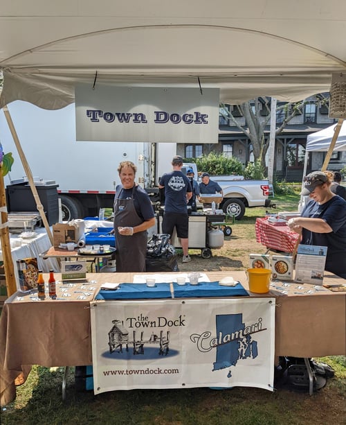 Under the large event tent, The Town Dock's table with sample cups and grill in the background