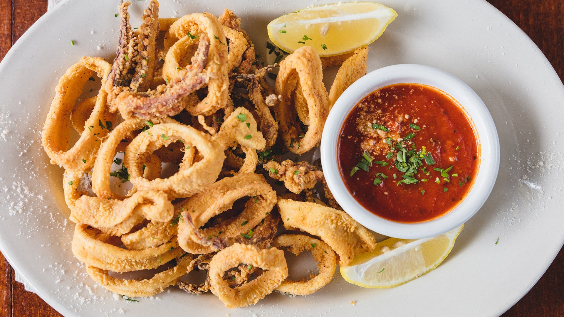 A white plate of fried calamari rings and tentacles, with lemon wedges, a side of marinara, and dusting of parmesan cheese