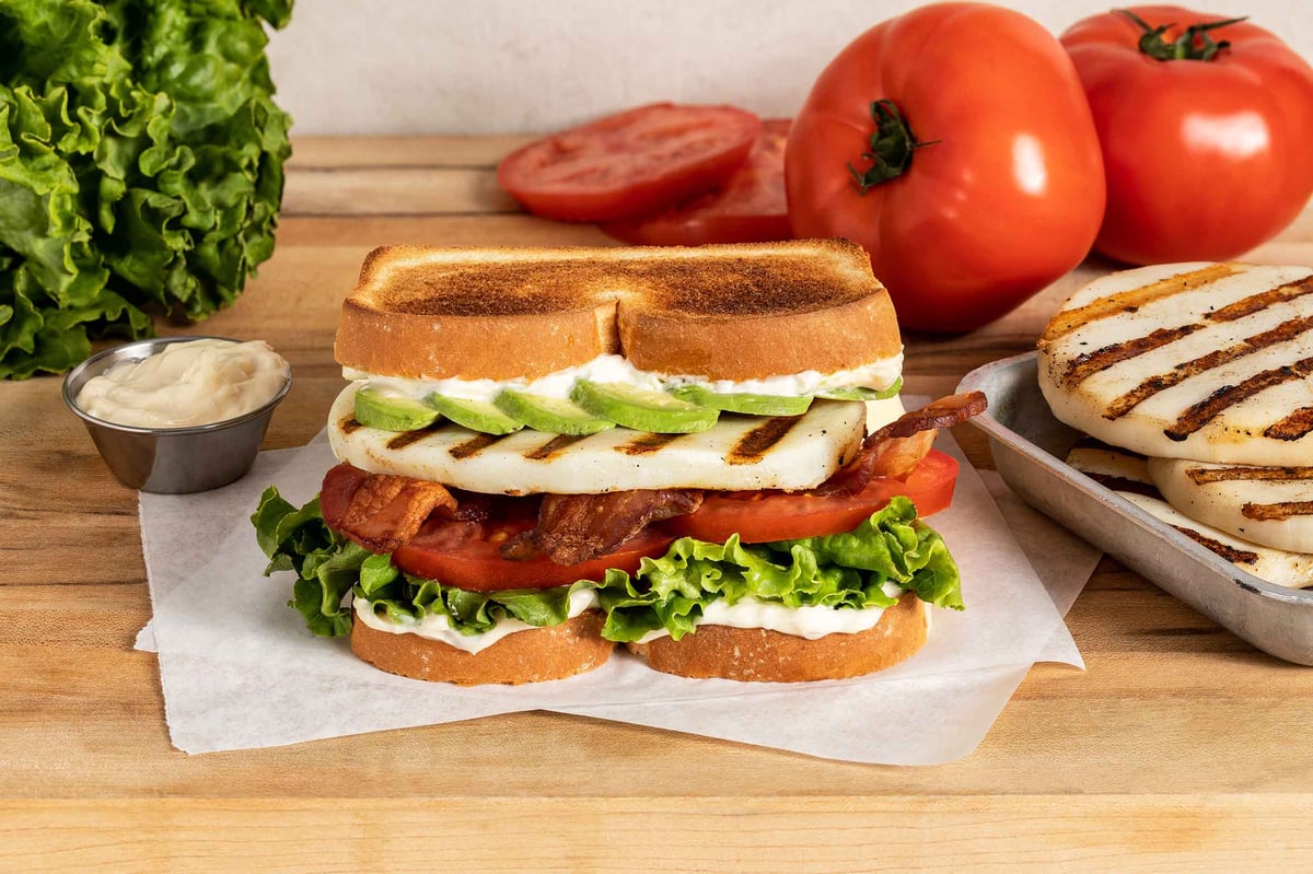 A hero image of our Smoked Calamari Avocado BLT: layers of mayonnaise, lettuce, tomato, bacon, a smoked and grilled calamari steak, avocado, and more mayo on thick, toasted white bread.