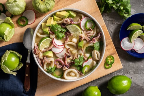 A white bowl full of calamari rings and tentacles, radishes, limes, jalapenos, avocado, and cilantro. It sits on a pale wood cutting board on a concrete counter with tomatillos decorating.