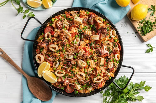 A black paella dish filled with calamari rings, peas, rice, and chorizo sits on a white background with lemons