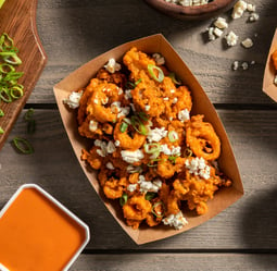 A kraft paper bowl with fried calamari rings tossed in buffalo sauce, garnished with slice scallions and blue cheese. Perfect for the big game!