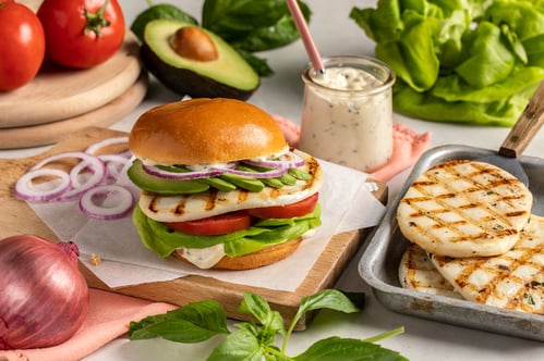 A grilled calamari steak stacked with red onion rings, avocado, tomato, lettuce, and sauce on a brioche bun. 