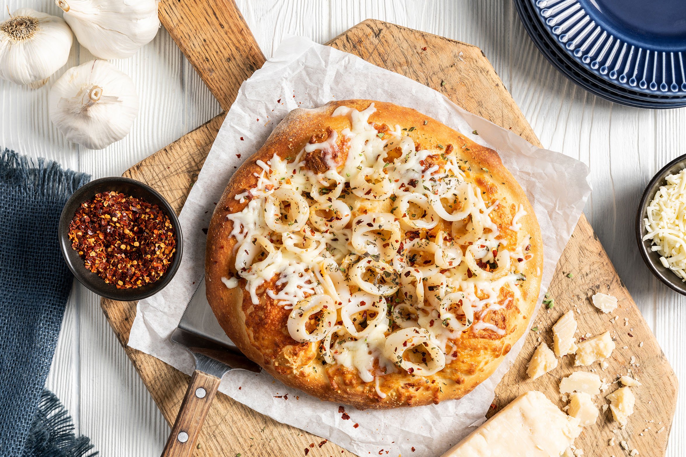 A pizza on a wooden pizza peel. The pizza has a medium-thick crust, no red sauce, melty cheese, and calamari rings topped with crushed red pepper and oregano garnish. 