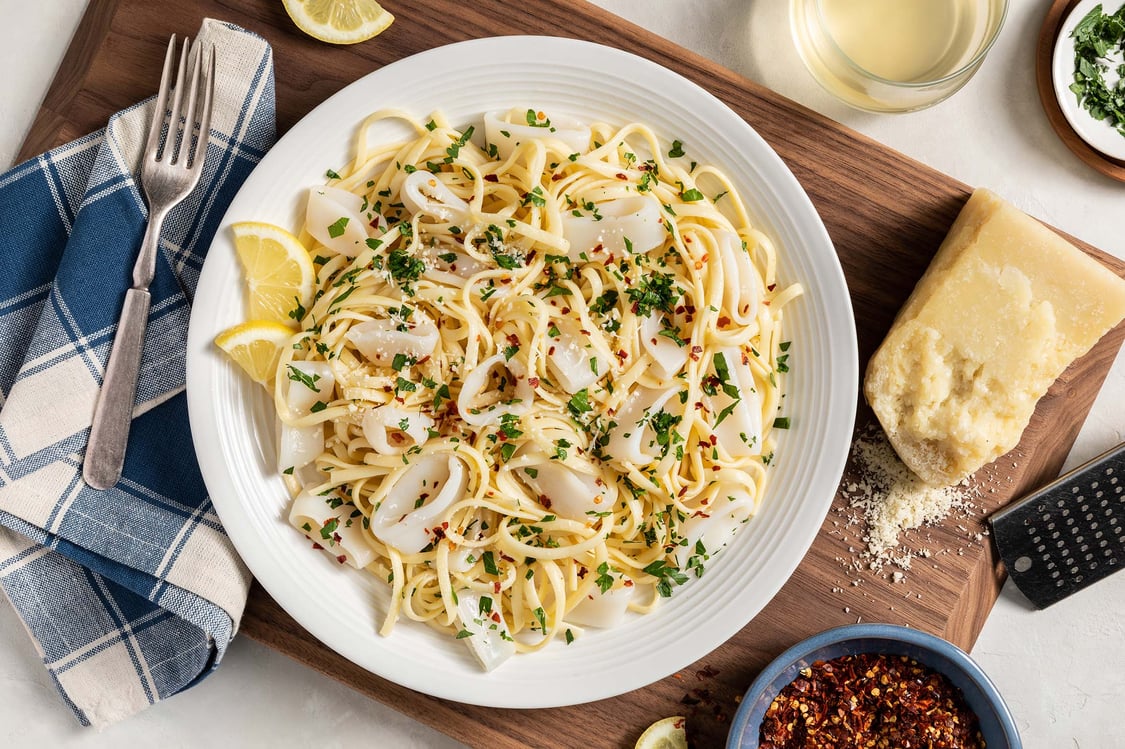 A round white dish of linguine and calamari rings, topped with parmesan, parsley, and red pepper flakes. Scattered around the dish are smaller bowls of the ingredients and a blue plaid napkin with a fork.