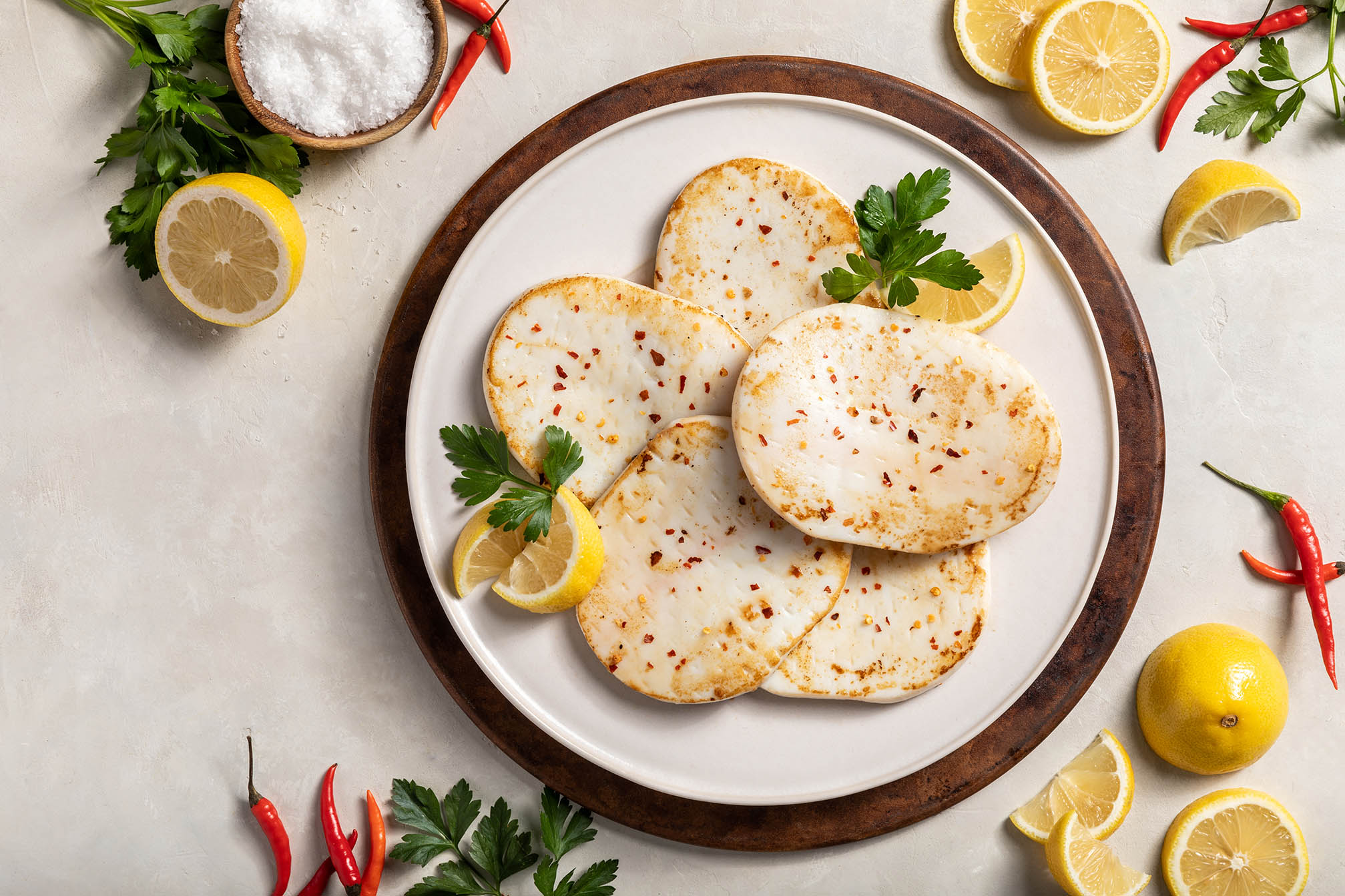 A plate of lightly cooked calamari steaks, garnished with parsley, lemons, and small hot peppers